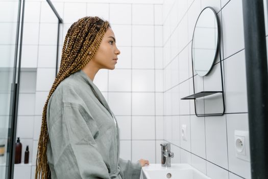 Side profile of a woman with braids looking at a mirror in a minimalist bathroom.