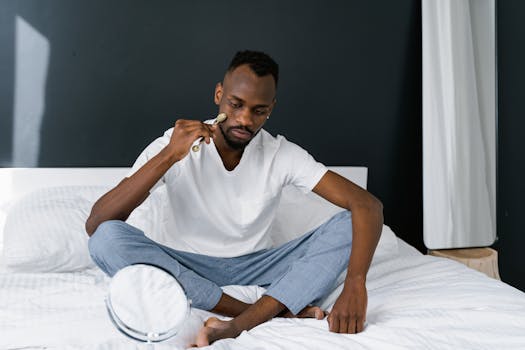 Black man sitting on bed, using a jade roller for skincare in a cozy bedroom setting.
