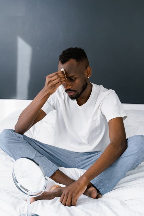 Free A man in a white shirt sits on a bed, focusing on self-care with a mirror. Stock Photo