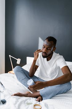 A man indulges in self-care by applying facial cream while sitting on a bed.