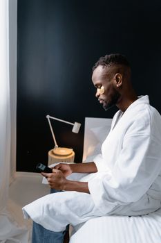 A man wearing a bathrobe relaxes in a bedroom while using a mobile phone for self-care.