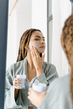 Woman in a green robe applying facial cream in front of the mirror for skin care.