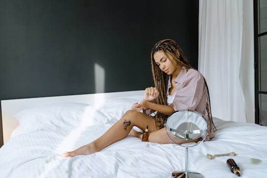 Woman with tattoos and braided hair applying lotion, sitting in a modern bedroom.