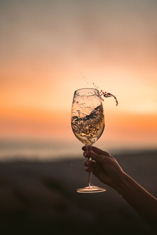 Free Hand holding a splashing wine glass against a vibrant sunset beach backdrop. Stock Photo