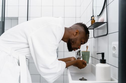 Free Young man in bathrobe washing face in a modern white bathroom. Stock Photo