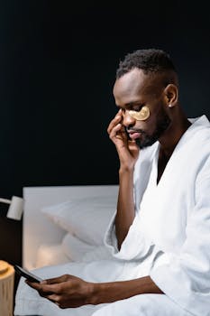 African American man in bathrobe applies under eye masks while using a smartphone indoors.