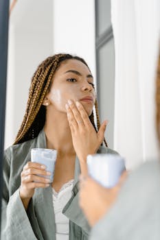 A woman with braided hair applying facial cream indoors, focusing on skincare routine.
