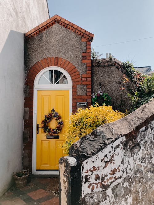 Free Old stone residential house with bright yellow door decorated with floral wreath against cloudless blue sky in town Stock Photo