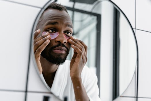 Man in white bathrobe applying eye patches in bathroom mirror for skincare routine.