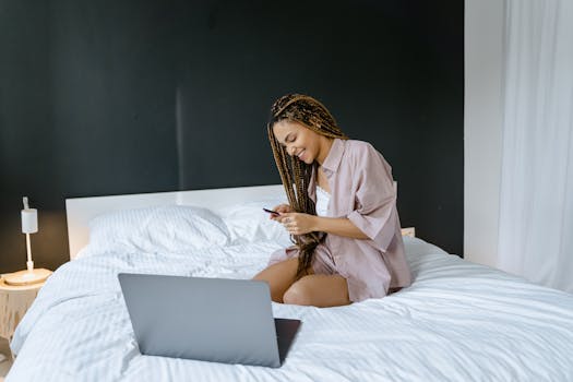 Woman with braided hair using smartphone on bed with laptop in cozy setting.