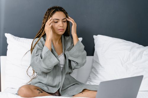 Free A young woman with braided hair sits in her bedroom, using a laptop, deep in thought. Stock Photo