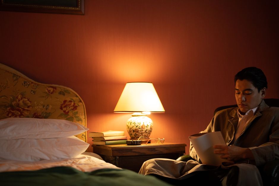 A man sits in a cozy hotel room reading under warm, ambient light.
