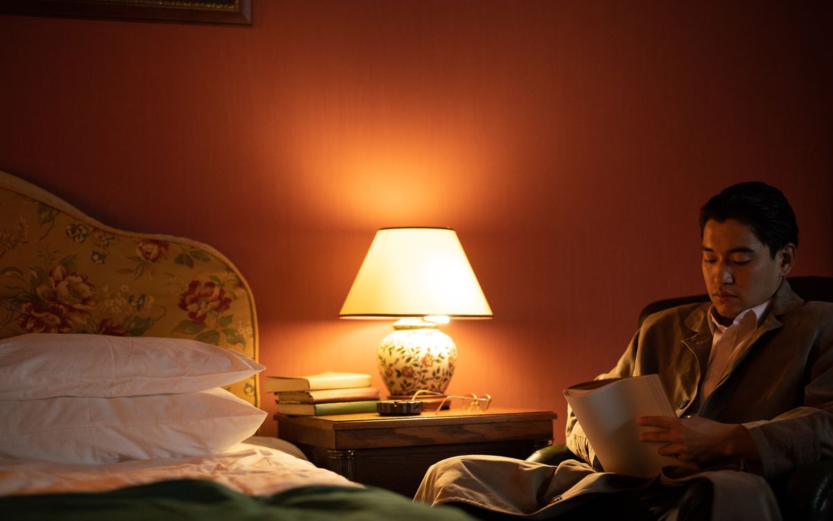 A man sits in a cozy hotel room reading under warm, ambient light.