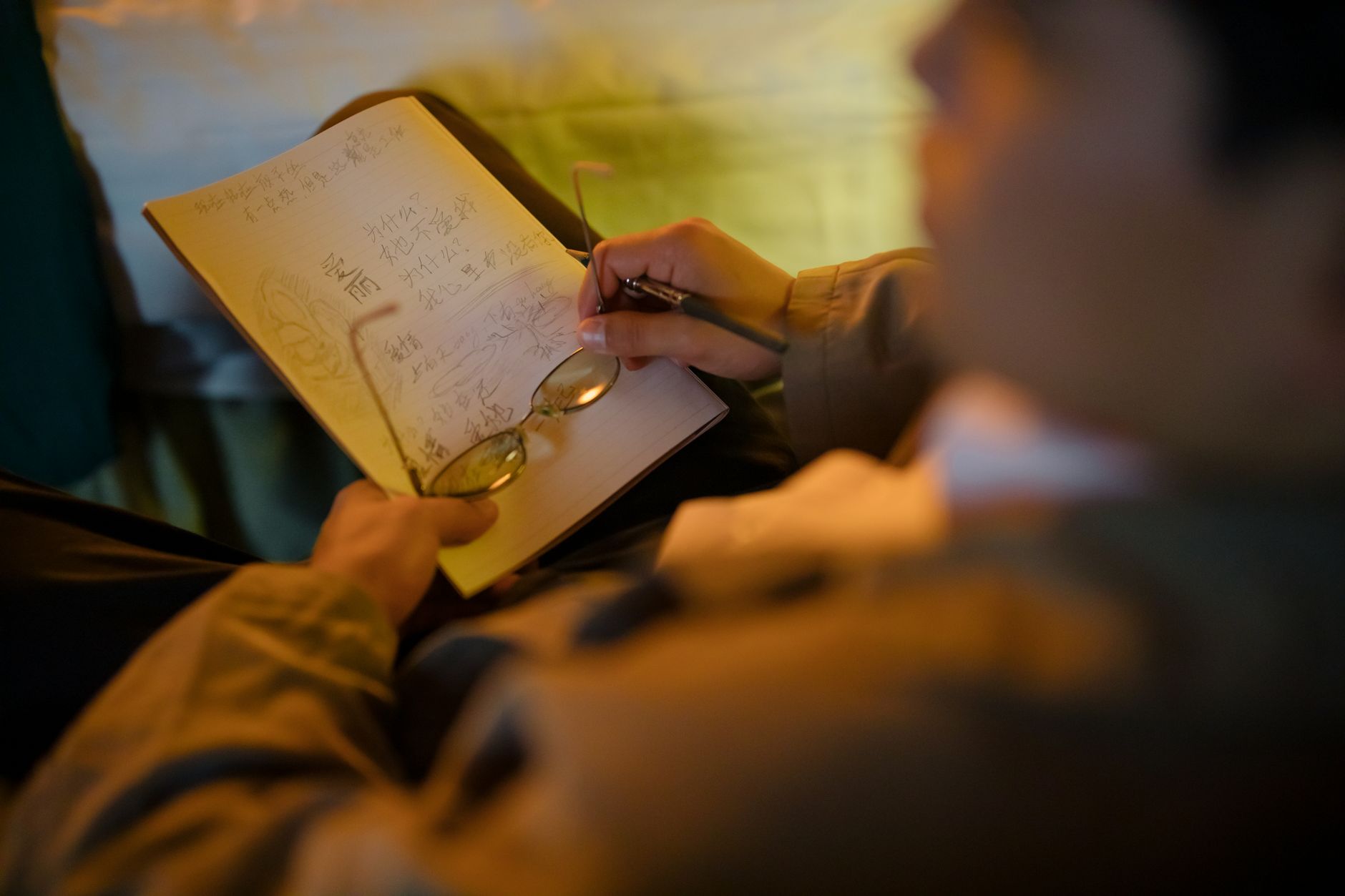 A man is writing in a notebook with glasses in a dimly lit environment, focused on his task.