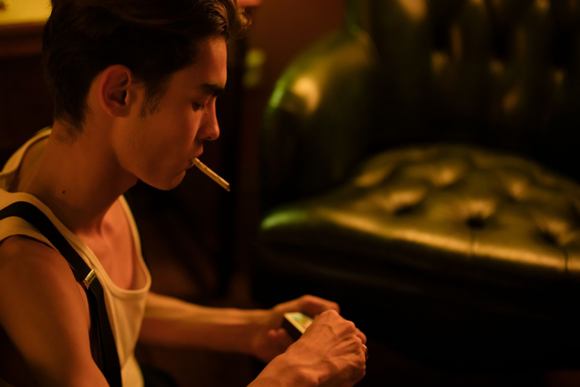 A young man sits indoors, smoking a cigarette in a dimly lit room.