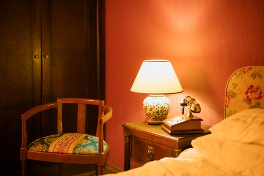 Warm vintage bedroom interior featuring a lamp, chair, and telephone by the bedside.