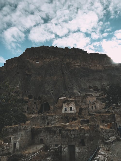 Ancient derelict stone buildings located near tall rough mountain against blue sky with clouds in rural area on summer day