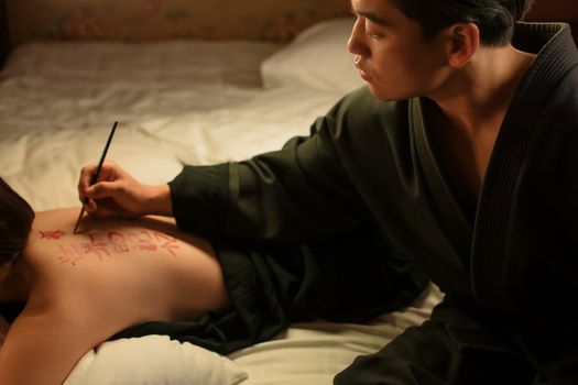 Male artist creating intricate calligraphy art on woman's back indoors, showcasing concentration and tradition.