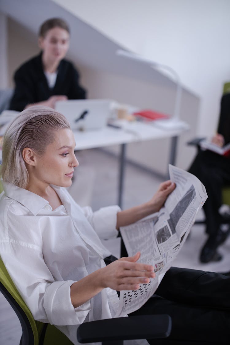 A Woman In White Long Sleeves Sitting On A Chair While Reading Newspaper