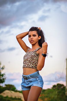Confident young woman with curly hair posing outdoors in summer wearing a printed top and denim shorts.