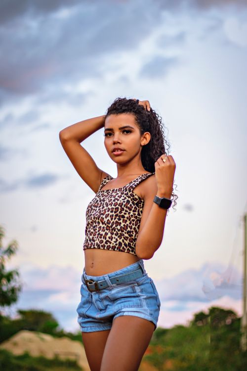 Free Confident young woman with curly hair posing outdoors in summer wearing a printed top and denim shorts. Stock Photo
