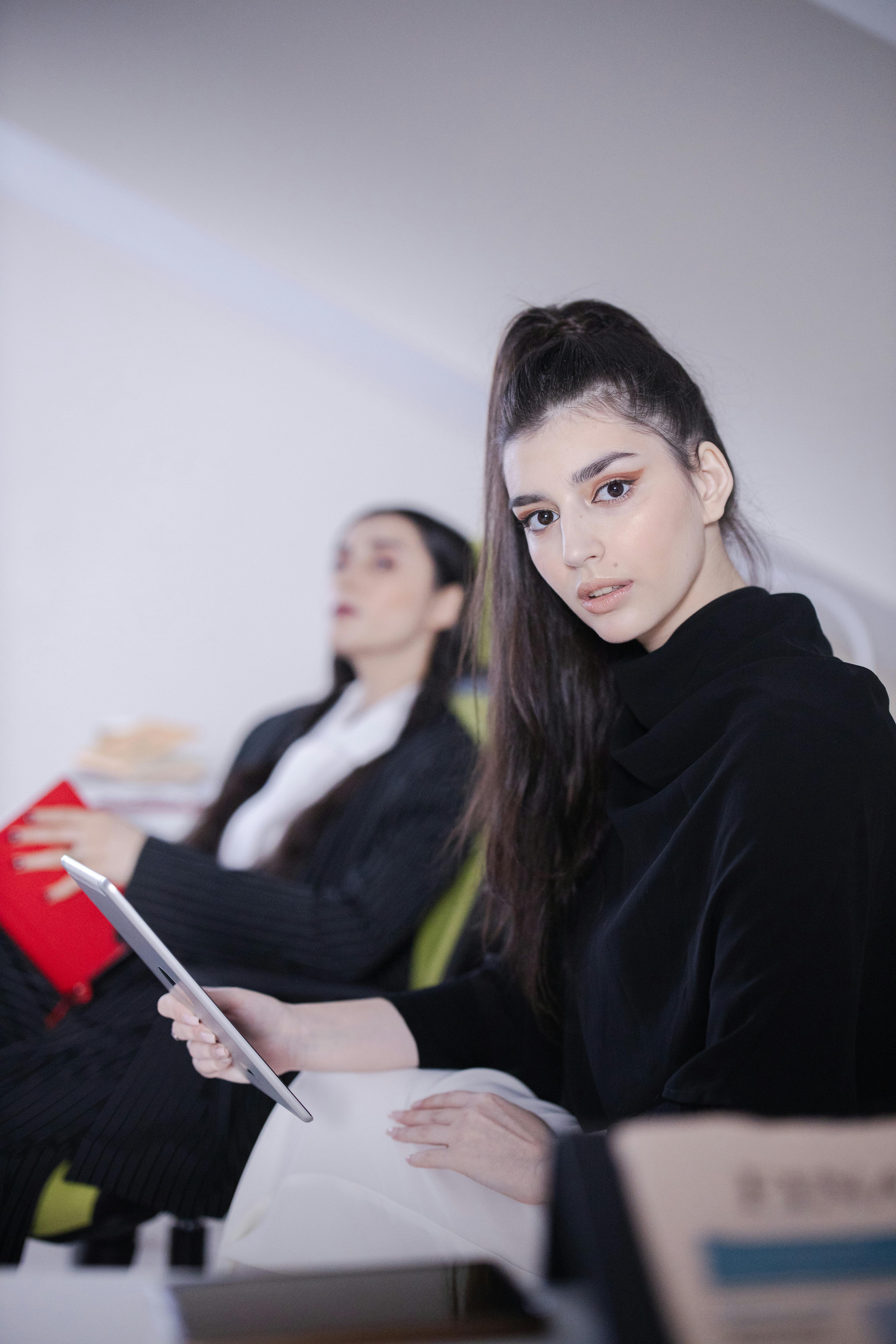 Two professional women in a modern office setting engaged with technology and reading materials.