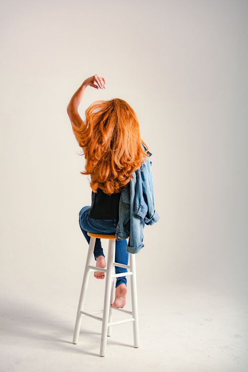 Free Back view of anonymous barefoot female teenager with long red hair and denim jacket sitting with raised arm on light background Stock Photo