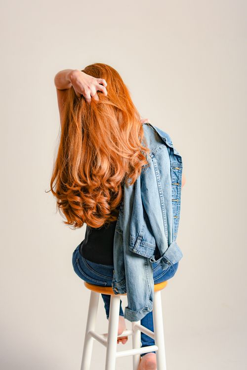 Free Back view of anonymous trendy woman with denim jacket touching long hair while sitting on light background Stock Photo