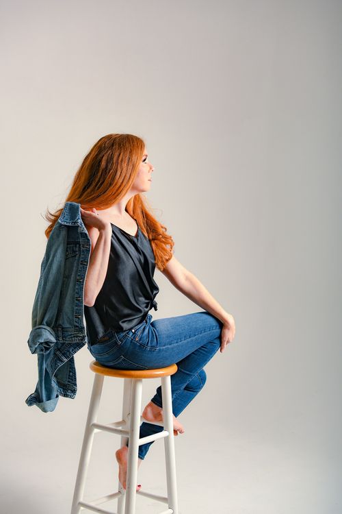 Free Side view full body of barefoot self assured female model wearing denim jeans and silk blouse sitting in studio Stock Photo