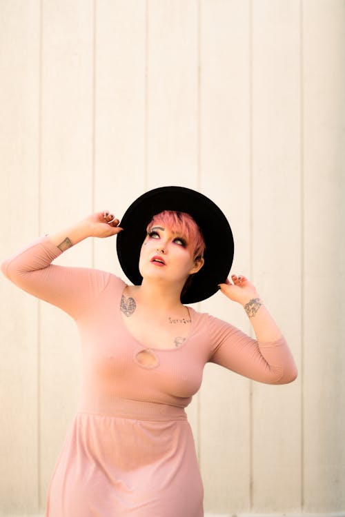 Stylish young female with pink hair wearing stylish hat and dress looking up with mouth opened on white background