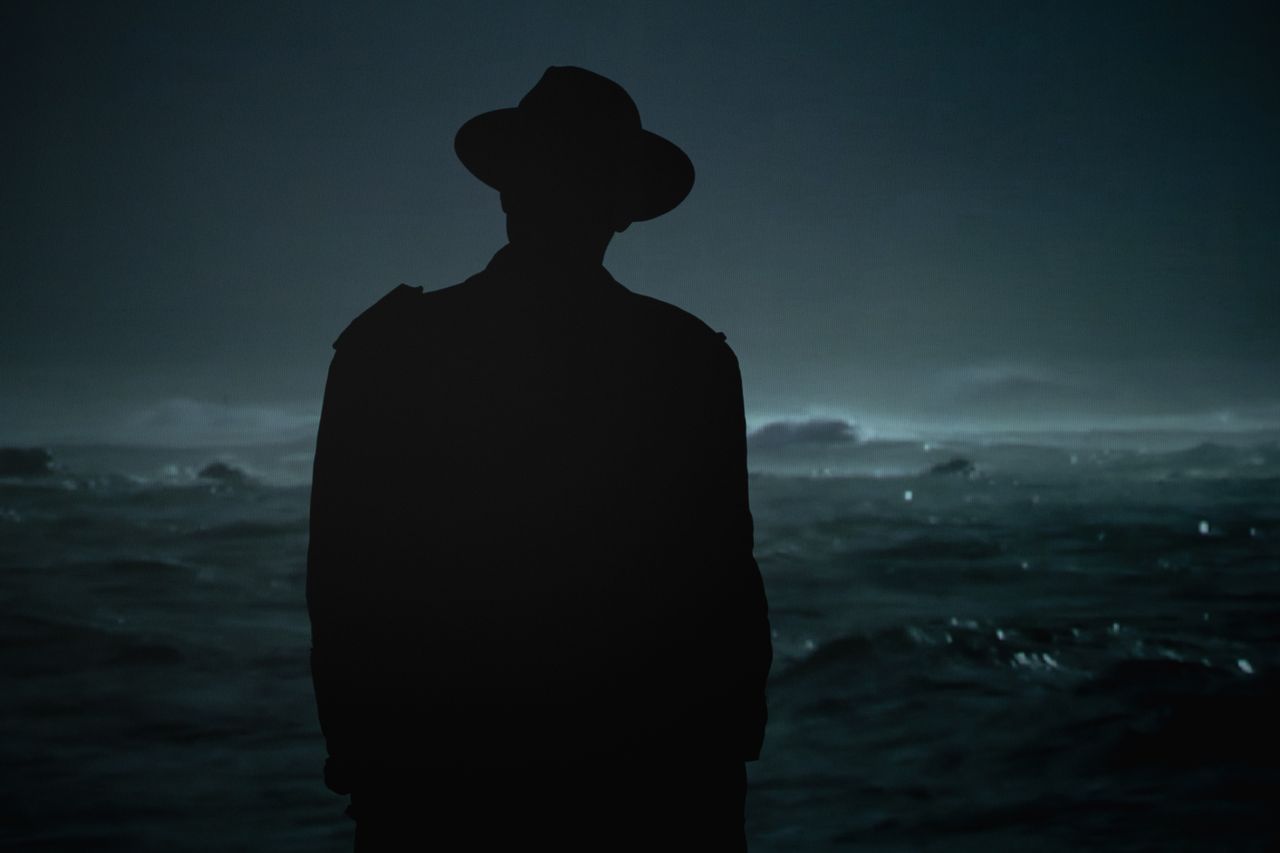 dramatic silhouette of a man with hat standing by the ocean under a da