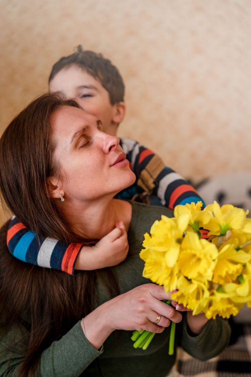 Free Mother and son sharing a joyful hug with bright yellow flowers indoors. Stock Photo