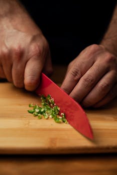 Close-up of hands slicing green peppers on a wooden chopping board with a red knife.