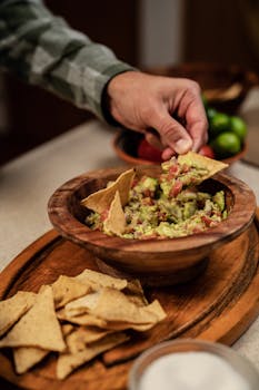 Close-up of a hand dipping tortilla chips into fresh guacamole on a wooden tray.