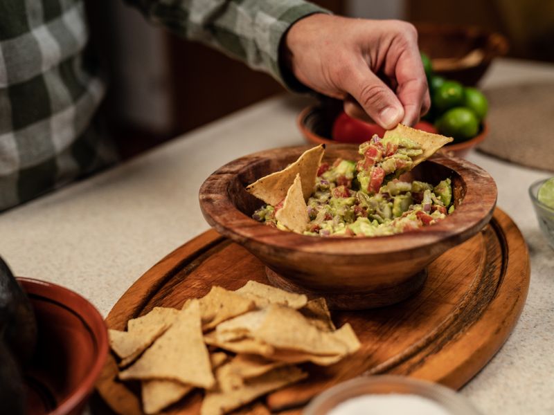 A hand dips a tortilla chip into a bowl of fresh guacamole with tomatoes and limes.