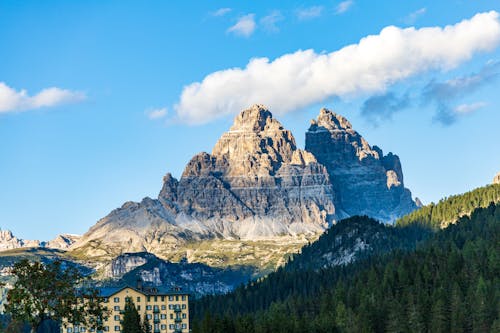 Picturesque scenery building placed near green hills covered with coniferous forest against Dolomite Mountains under cloudy blue sky in sunny day