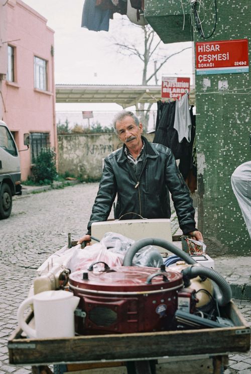 Free A man in a black leather jacket pushes a cart filled with various items on a cobblestone street. Stock Photo