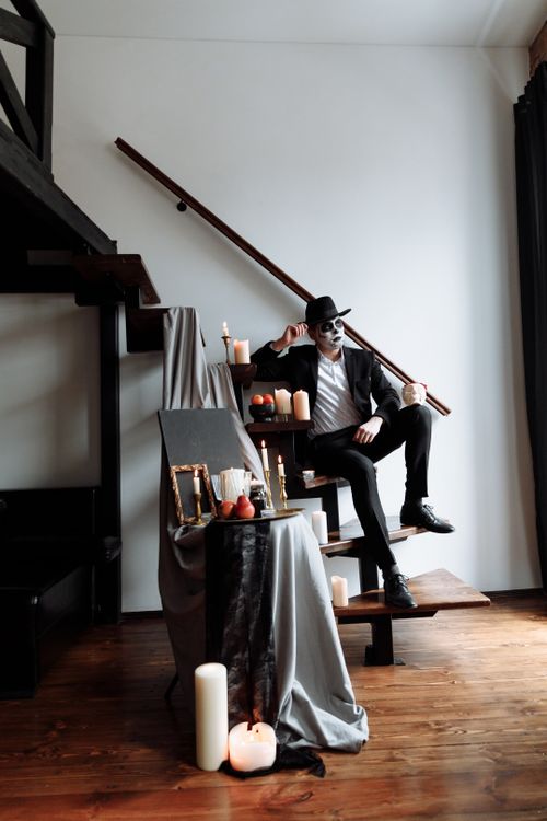 Free A man in artistic Day of the Dead makeup and black suit sits on wooden stairs with candles. Stock Photo