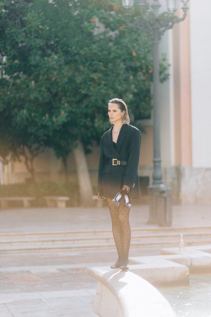Fashionable woman in a black dress standing near a fountain on a sunny day outdoors.