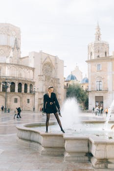 A fashionable woman poses by a water fountain in a historic European square, exuding style and elegance.