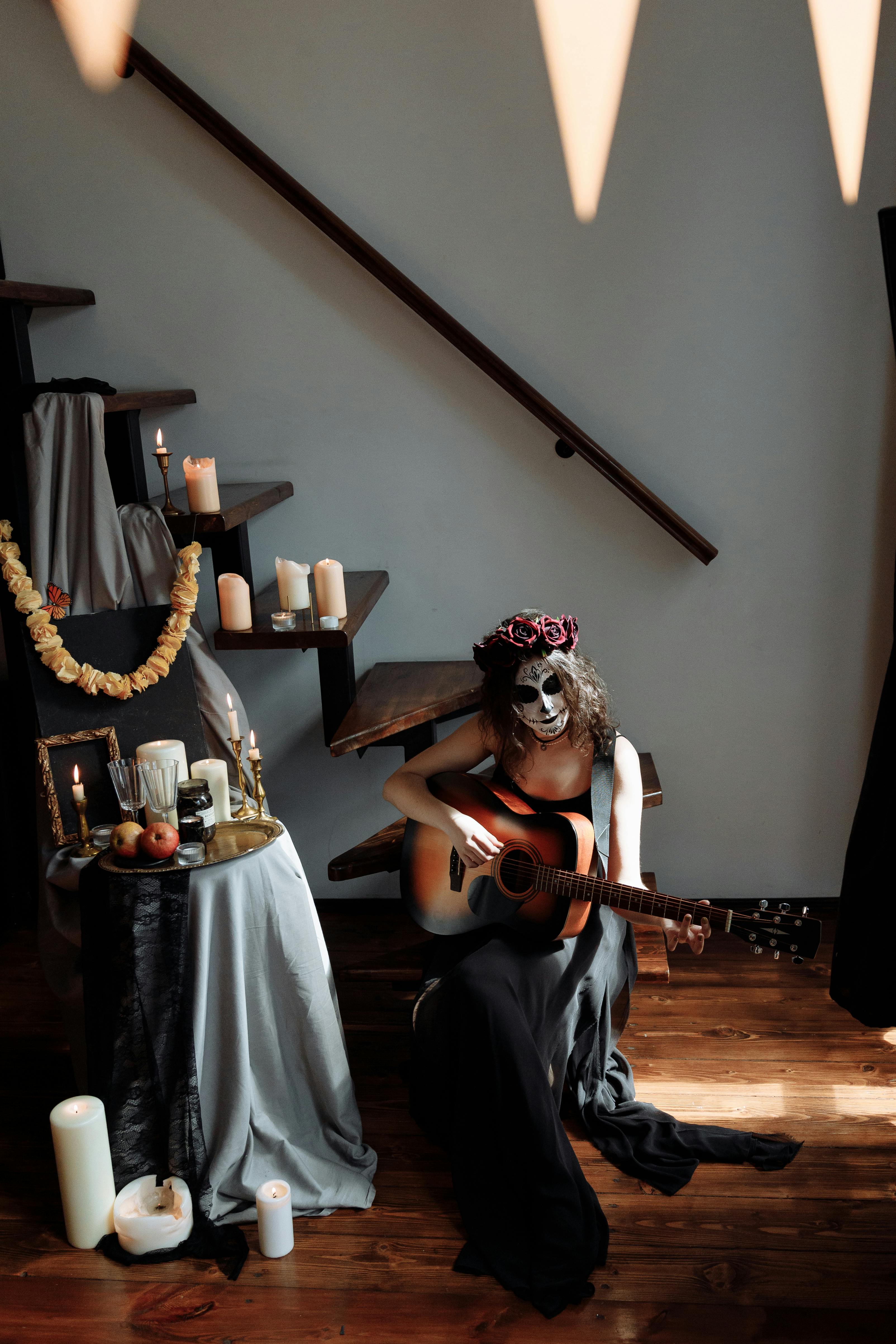Woman in Dia de los Muertos makeup playing guitar beside an altar indoors.