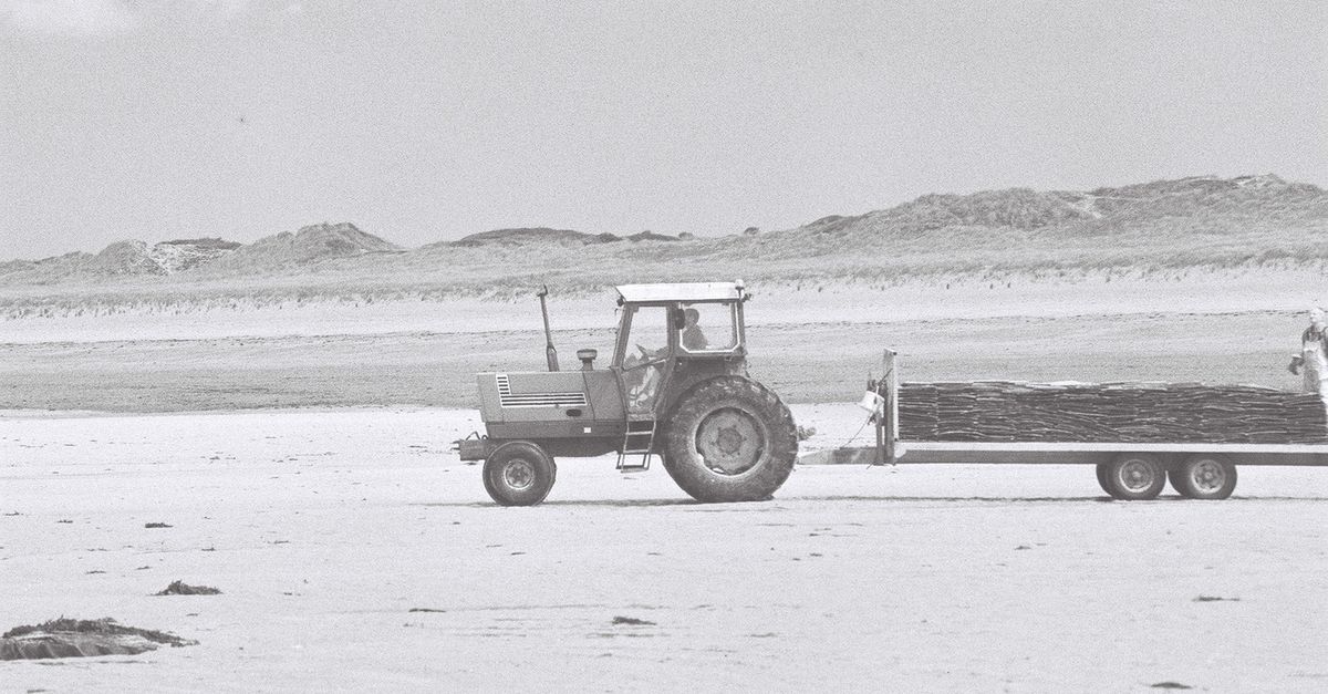 Black and white photo of a tractor towing a trailer on a sandy beach.