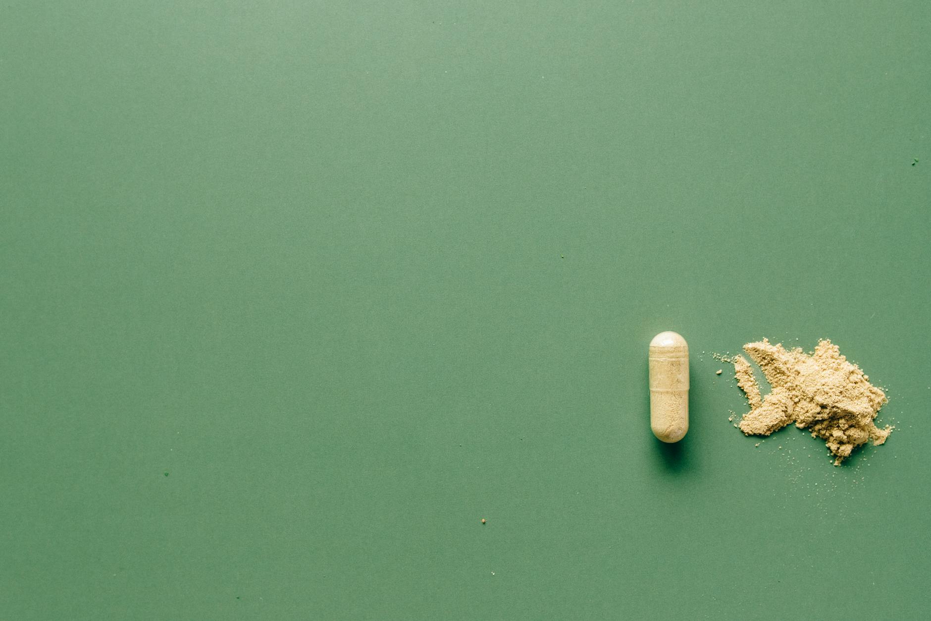 Close-up of herbal supplement capsule and powder on a minimalist green surface.