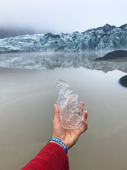 Hand Holding a Piece of Ice with Lake and Snowcapped Mountains in the Distance 