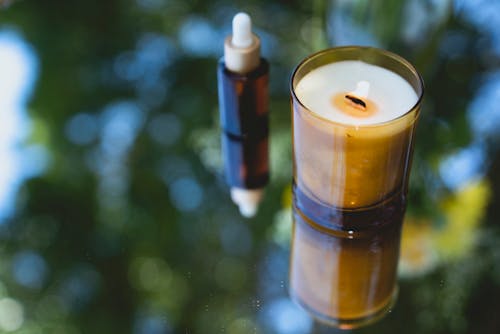 High angle of fragrant burning candle placed near amber glass bottle of essential oil on reflecting table