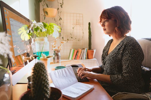 A woman focused on work at her home office setup with plants and a desktop computer.