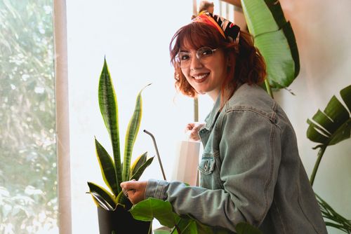 Free Smiling woman in denim jacket watering houseplants indoors with natural light. Stock Photo
