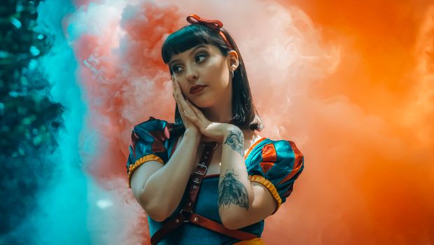 Portrait of a tattooed woman in a Snow White costume amid vibrant smoke clouds. Conceptual and artistic.