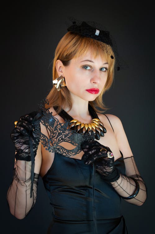 Charming young red hair female wearing elegant black dress and gloves and holding black carnival mask while looking at camera against black background