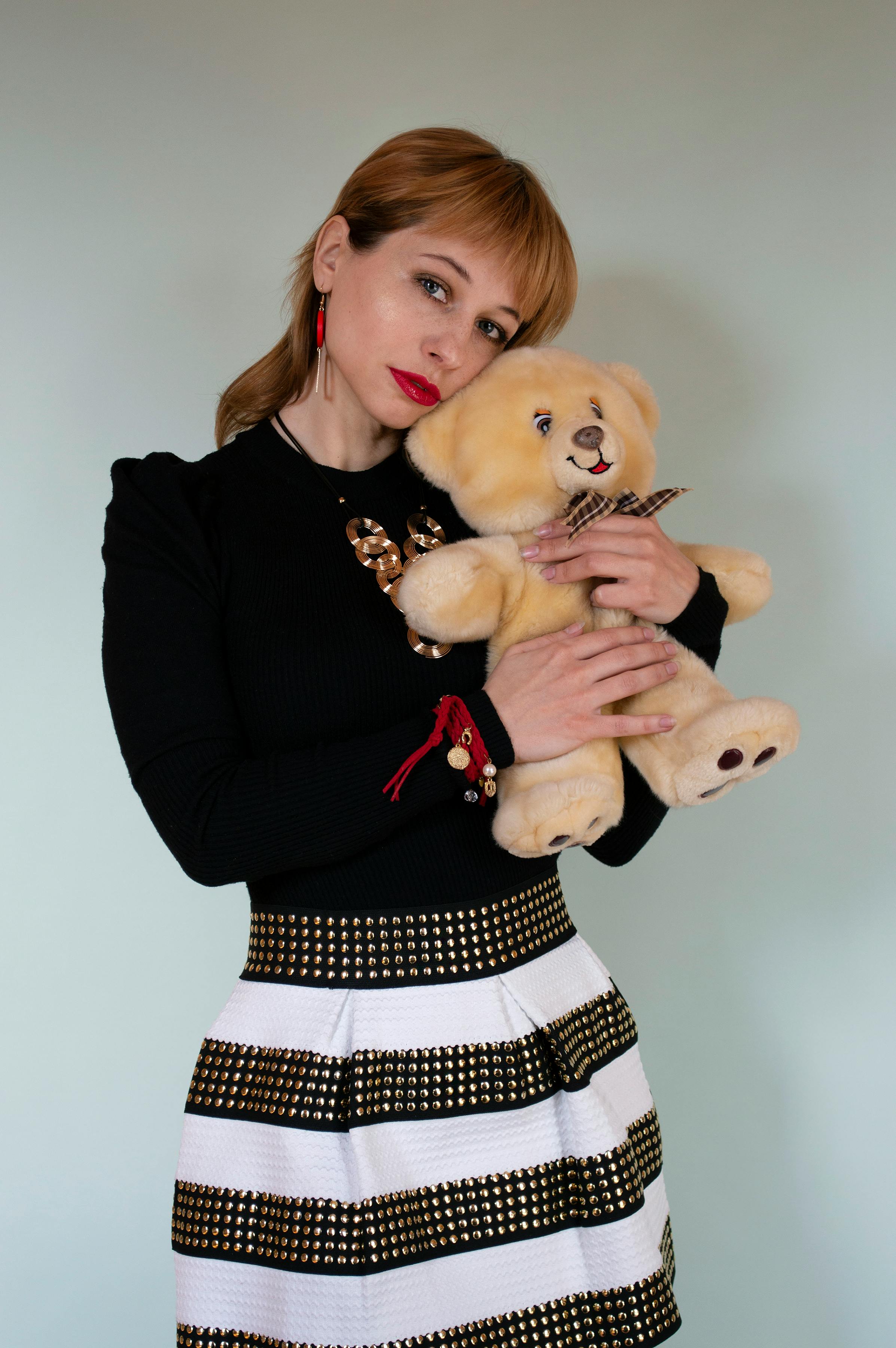 Young red hair female wearing mini skirt and black long sleeve shirt embracing toy bear while looking at camera against white background